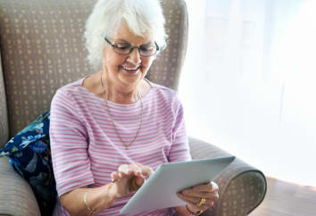 White older woman smiling, sitting in an armchair, using a tablet